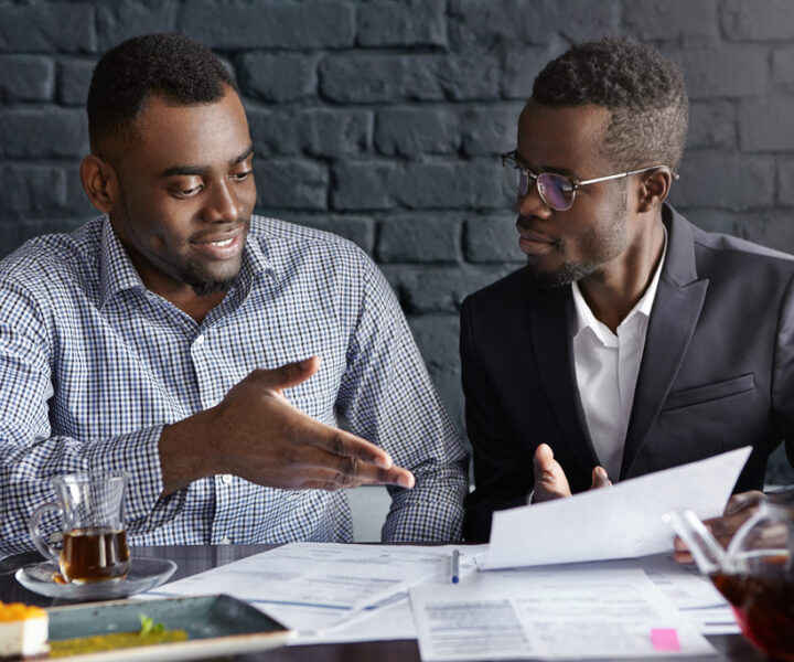 Attractive African businessman in glasses and suit holding papers, showing presentation of his project to cheerful business partner, who smiling happily, supporting his idea with positive gesture